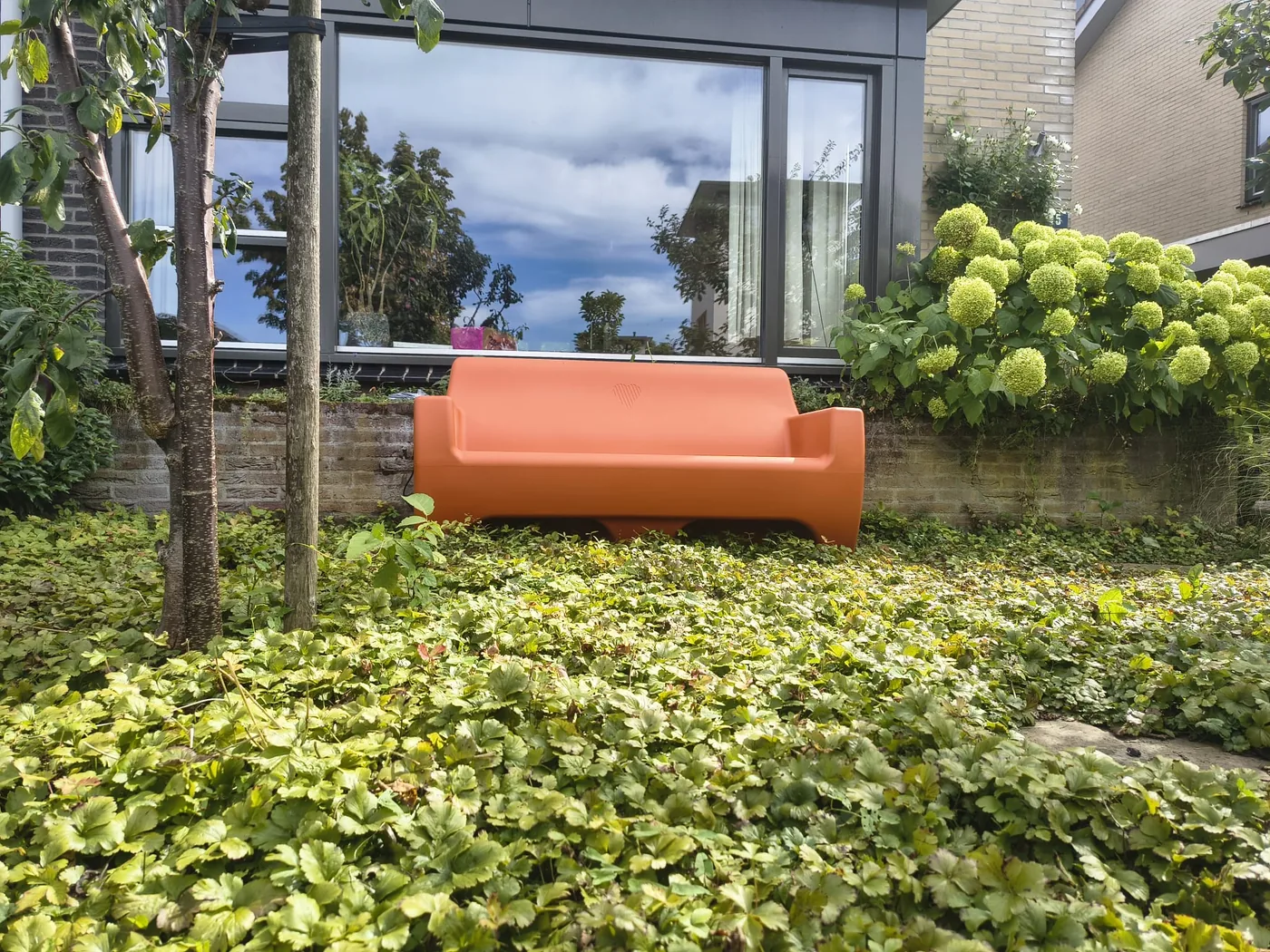 Papaya oranje HappySofa regenbank in een voortuin, met bodembedekkers ervoor en hortensia's erachter